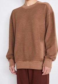 Sudadera marrón con un acabado texturizado, puños y dobladillo acanalados. Presenta un logo sutil y un detalle de bolsillo lateral.