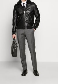 Schwarze Lederbomberjacke mit Pelzkragen, Reißverschluss vorne, gerippten Bündchen und einer grauen Hose, hält eine schwarze Tasche.