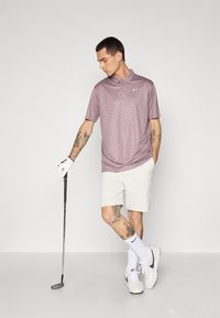 Polo s krátkym rukávom, s golierom, svetlofialovej farby s čiernymi bodkami, v kombinácii s bielymi šortkami. Doplnky zahŕňajú golfovú rukavicu a topánky.