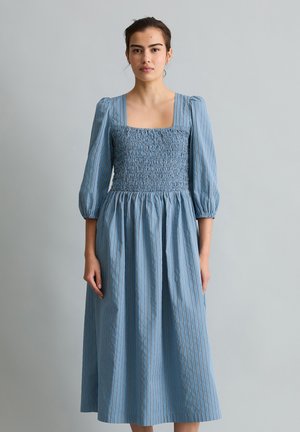 Frau trägt ein blau gestreiftes smockiertes Midikleid mit quadratischem Ausschnitt und gepufften Dreiviertelärmeln, steht vor einem einfarbigen grauen Hintergrund.