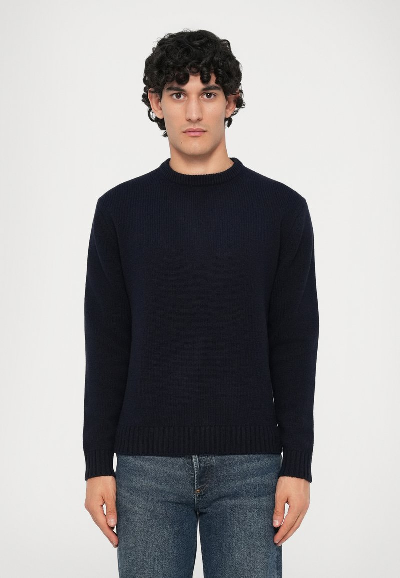 Uomo con capelli ricci che indossa un maglione blu scuro semplice e jeans blu, in piedi contro uno sfondo grigio chiaro.