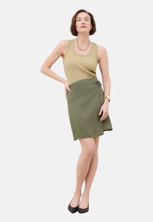 Femme debout, une main sur la hanche, portant un haut en maille beige sans manches, une jupe portefeuille vert olive avec boucle, et des chaussures noires pointues.