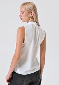Blusa bianca senza maniche con un colletto alto, chiusura a bottoni sul retro, texture liscia e orlo curvo, abbinata a jeans scuri.