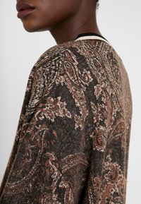 Pull en laine marron à motif paisley complexe, avec une texture douce et un col en rib blanc et noir. Vue de côté en gros plan.