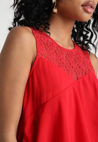 Blouse rouge sans manches avec une insertion en dentelle au niveau de l'encolure, silhouette fluide et tissu léger et lisse.