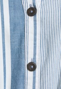 Pruhovaná košile v modro-bílé látce, která má zapínání na knoflík s tmavě hnědým knoflíkem a kontrastním prošíváním podél légy.