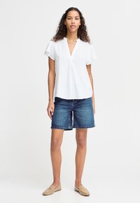 Femeie cu păr creț care poartă o bluză albă cu mâneci scurte și textură, pantaloni scurți din denim până la genunchi, mocasini bej și un colier cu lanț auriu.