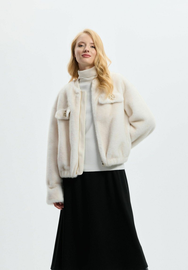 Chaqueta de piel sintética en color crema, con cierre de cremallera, dos bolsillos delanteros y puños acanalados. Llevada sobre un suéter de punto blanco con cuello alto y una falda negra.