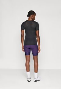 Sort i svart mesh med korte ermer, parret med purpur fargede kompresjonsshorts med teksturerte detaljer. Modellen har på seg hvite sokker og sportssko.