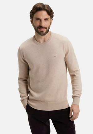 Pull beige à col rond en matière douce, avec des poignets et un ourlet côtelés, arborant un petit logo violet sur la poitrine. Porté avec un pantalon foncé.