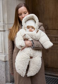 Krämfärgad fluffig fleecebabyoverall med en stor huva och träknappar. Barnet har blå ögon och bär en vit stickad mössa.