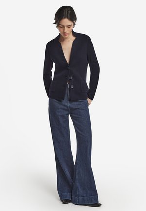 Marineblauer Cardigan aus strukturiertem Strick mit tiefem V-Ausschnitt, drei Knöpfen und zwei vorderen Taschen, kombiniert mit weit geschnittenen Jeanshosen.