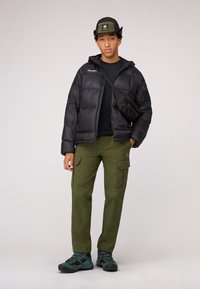 Doudoune noire avec capuche, pantalon cargo vert olive, t-shirt noir, associée à un chapeau seau vert et des chaussures de randonnée sombres.