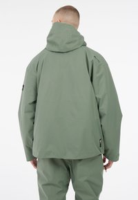 Zelena kapuljača, širokog kroja, glatke teksture, vodootporna tkanina, s diskretnim brandingom na ramenu i vezicom na vratu.