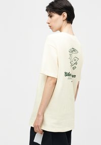 Γυναίκα με κοντά σκούρα μαλλιά, που φοράει μια υπερμεγέθη κρεμ μπλούζα με πράσινο γραφικό ενός ατόμου που διαβάζει κάτω από μια ομπρέλα στην πλάτη.