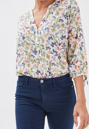 Femme portant une blouse blanche à fleurs à boutons avec des manches trois-quarts, associée à un jean bleu foncé taille haute.