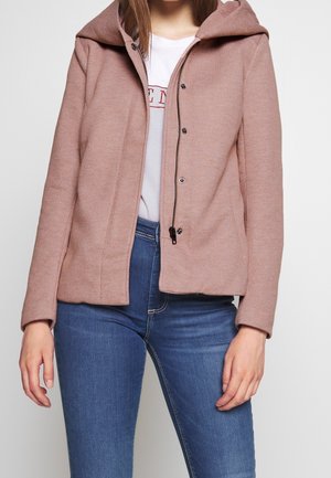 Femme portant une veste à capuche rose sur un t-shirt blanc graphique et un jean bleu, debout devant un fond uni clair.