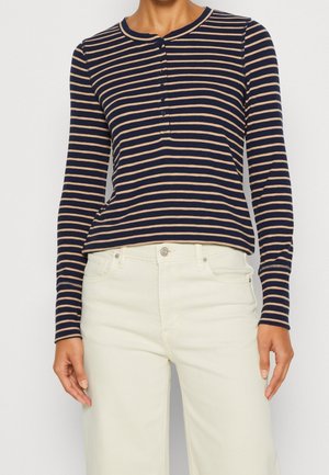 Femme portant un haut henley à manches longues à rayures horizontales bleu marine et beige, associé à un pantalon taille haute blanc cassé.