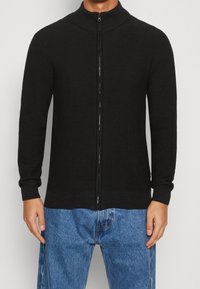 Cardigan noir en maille avec fermeture éclair, col montant, manches longues, poignets et ourlet côtelés; associé à un jean en denim bleu.