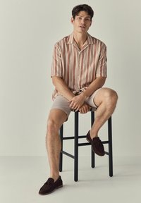 Kurzärmliges Hemd mit Knopfleiste und gestreiftem Muster in verschiedenen Rosa- und Cremetönen, kombiniert mit beigen Shorts und dunkelbraunen, geflochtenen Loafers.