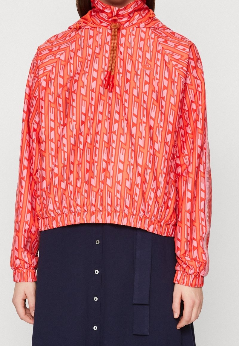 Femme portant un pull rouge vif à motifs avec poignets et ourlet élastiques, associé à une jupe foncée boutonnée.
