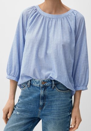 Vrouw draagt een loszittende, lichtblauwe blouse met lange mouwen en subtiele strepen, gecombineerd met een high-waisted blauwe spijkerbroek.