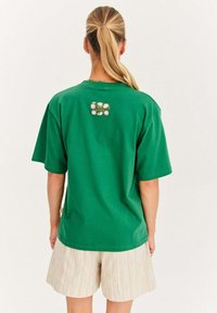 Zelené bavlnené tričko s krátkymi rukávmi, s kvetinovým vzorom na tagu vzadu. V kombinácii s pásikavými béžovými šortkami.