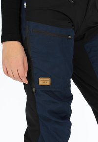Marineblauwe en zwarte outdoorbroek met een gestructureerde afwerking, ritszakken en een tan label met "SWEDEMOUNT" aan de zijkant.