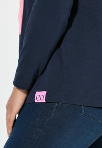 Camiseta de manga larga en color marino con una etiqueta rosa en el dobladillo, que presenta una textura suave y un diseño sencillo. Se lleva con jeans oscuros.