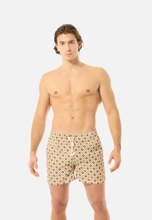 Bar overkrop mand med lys hud og brunt hår iført beige og hvide badebukser med diamantmønster og snor, stående mod en ensfarvet baggrund.