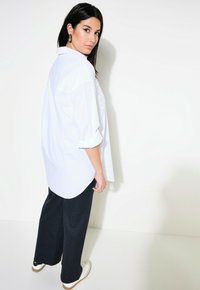 Camicia bianca larga con maniche arrotolate, abbinata a pantaloni neri a righe sottili e scarpe da ginnastica bianche. Insieme semplice e casual.