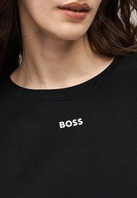 Κοντινό ενός ατόμου που φοράει μαύρο πουκάμισο με τη λέξη «BOSS» τυπωμένη σε λευκό κοντά στο ντεκολτέ.