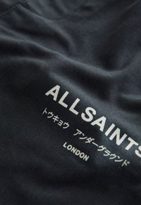 Čierna látka s textúrovaným povrchom, na ktorej je bielym písmom logo "ALLSAINTS", japonský text a nápis "LONDON" v dolnej časti.