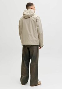 Veste imperméable beige clair avec capuche, coupe décontractée et tissu texturé ; associée à un pantalon en jean vert foncé ample.