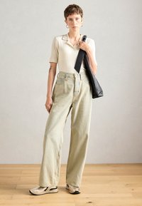 Une jeune personne aux cheveux courts se tient debout, vêtue d'un polo en crème à côtes, d'un jean large en denim clair, de baskets et d'un sac à épaule noir.