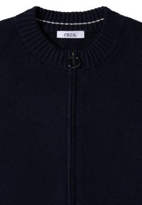 Námořnická modrá pletená kardigán s ribovaným límcem, má zipový uzávěr a kulatou černou táhlo, značkovou nálepku "CECIL" na krku.