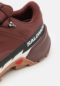 Brun atletikskon med texturerad tygöverdel, svart och vit branding, gummisula och framträdande orangea greppklackar.