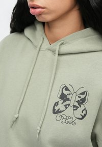 Egy nő világos zöld kapucnis pulóvert visel, amelyen lepkés grafika és az "OBEY" felirat látható a mellkasán, közelről mutatva a nyakát és az ajkait.