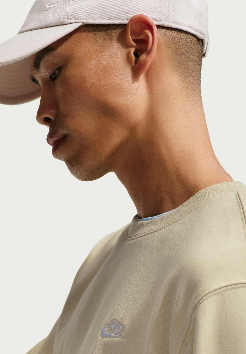 Sweat-shirt beige à col rond avec logo brodé, porté sous une casquette de couleur claire, montrant un profil mince et une texture de peau lisse.