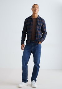 Flanellen overhemd in marineblauw en roestkleurige ruit met een knoopsluiting aan de voorkant en twee borstzakken, gecombineerd met blauwe spijkerbroeken en witte sneakers.