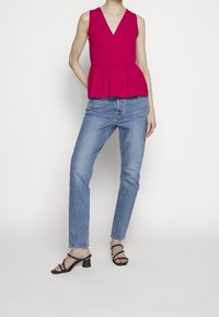 Femme portant un haut rouge sans manches à basque, un jean bleu et des sandales noires à talons, debout devant un fond blanc uni.