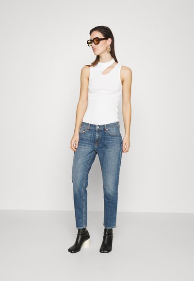 Hvit ermeløs topp med utskjæring, blå jeans og svarte ankelstøvler med hvite hælakse. Modellen har på seg oversized solbriller og øredobber.