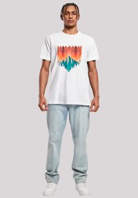 White cotton t-shirt featuring a multicolored graphic with mountains and trees. Worn with light blue denim jeans and white sneakers.
