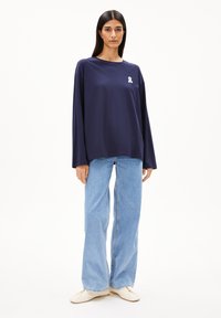 Marineblaue Langarmbluse aus Baumwolle mit einem weißen Band-Logo auf der Brust, kombiniert mit hellblauen weiten Jeans und beigen Schuhen.