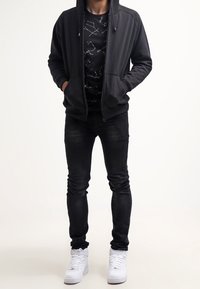 Black zip-up hoodie with front pockets, worn over a black t-shirt featuring white constellation patterns, paired with dark jeans and white sneakers.