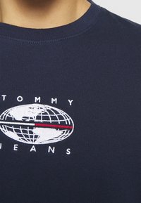 Mörkblå bomulls-T-shirt med en broderad jordglobdesign och texten "TOMMY JEANS", accentuerad av en röd horisontell linje. Rund hals.