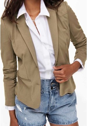 Personne portant un blazer beige sur une chemise blanche boutonnée, associée à un short en denim délavé bleu clair taille haute.
