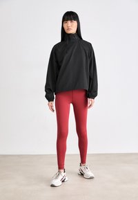 Schwarzer, leichter Pullover mit hohem Kragen und elastischen Bündchen, kombiniert mit roten Leggings und weißen Sneakern, die Akzente in Schwarz und Rot aufweisen.
