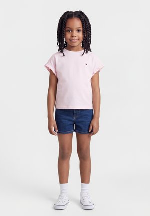 Young girl with braided hair wearing a light pink ruffle-sleeve shirt, dark denim shorts, white socks, and white sneakers, standing.