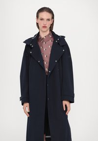 Tumesinine trench coat flared disainiga, suure kraega ja hõbedaste detailidega. Kandetav roosa mustriga särgi peal. Pikad varrukad nööpidega mansettidega.